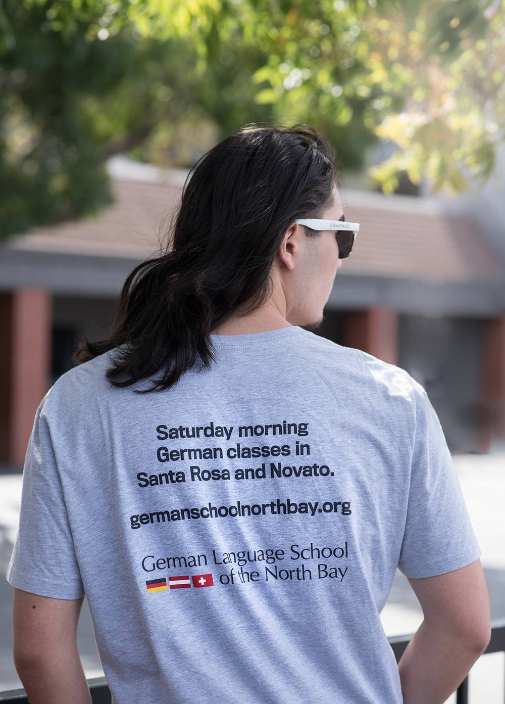 High school student posing with the back of school shirt that says "Saturday morning German classes in Santa Rosa and Novato." as well as the website and school logo.