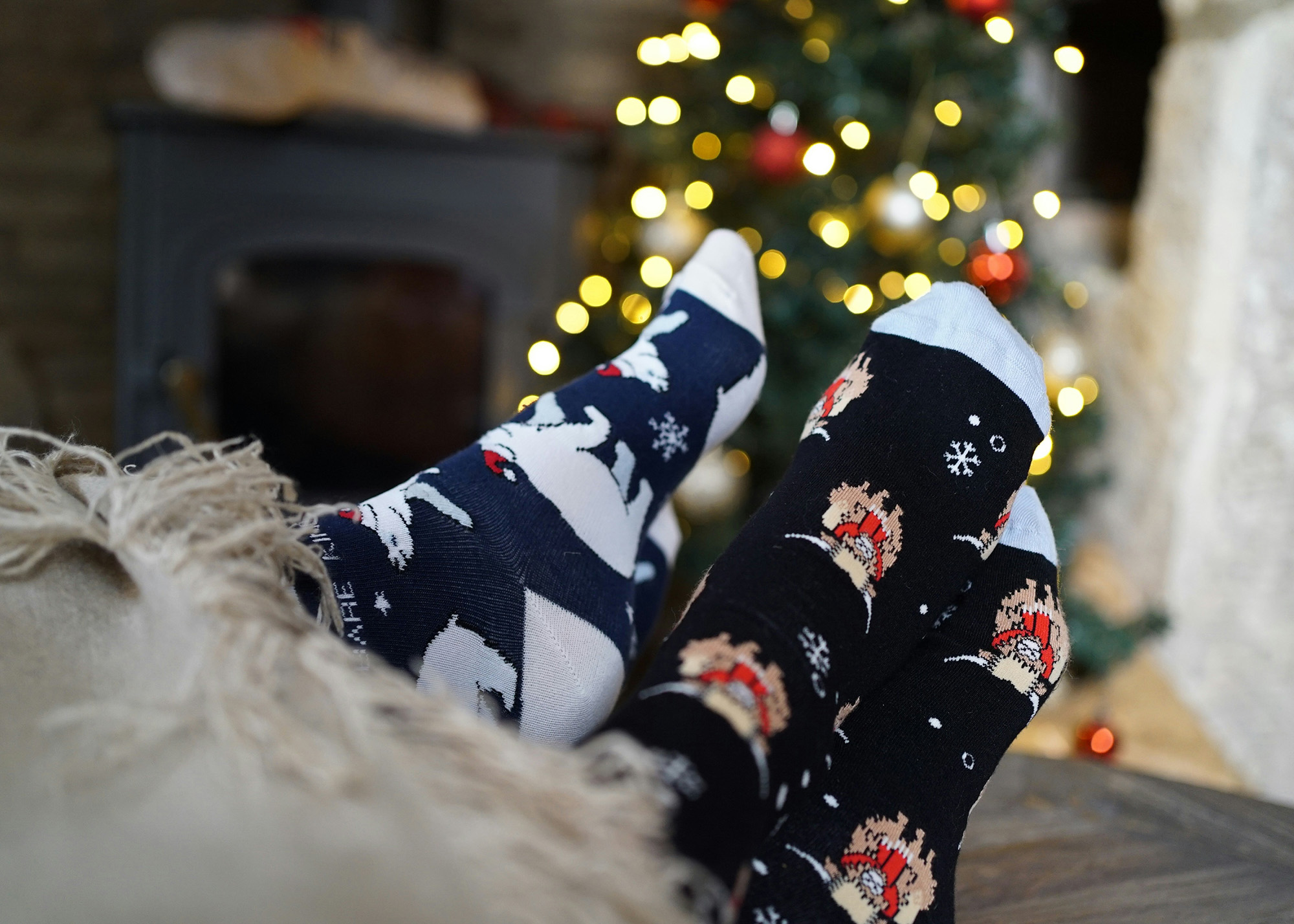 Two pairs of feet with winter-themed socks and a Christmas tree in the background.